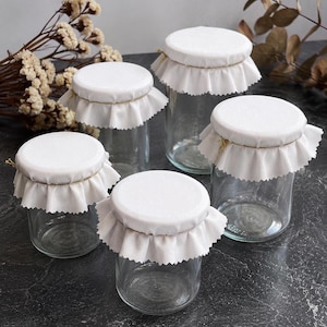 May include: A set of four clear glass jars with white fabric covers secured by thin gold twine. Each lid features a decorative ruffled fabric edge. The jars are empty and transparent, showcasing a simple cylindrical design with flat bases.