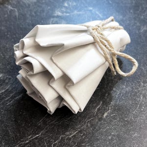 May include: A bundle of off-white fabric folded into multiple layers and secured with a piece of tan twine tied in a knot. The fabric has a smooth, slightly reflective surface and is arranged in a compact, gathered shape with visible folds.