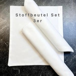Könnte beinhalten: Ein Set aus drei weißen Stoffbeuteln, von denen zwei aufgerollt und auf einem gefalteten quadratischen Beutel platziert sind. Der Text "Stoffbeutel Set 3er" ist auf dem Beutel aufgedruckt. Die Beutel liegen auf einer dunklen, strukturierten Oberfläche.