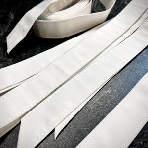 Könnte beinhalten: Nahaufnahme von weißen Stoffstreifen, einige flach und andere gerollt. Die Streifen haben eine strukturierte Optik und sind sauber vernäht. Der Hintergrund ist eine dunkle Oberfläche. Ideal für Näharbeiten.