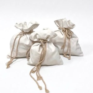 Può includere: Tre sacchetti con coulisse bianco sporco con lacci in corda beige. I sacchetti sono realizzati in un materiale morbido e strutturato e sono disposti su uno sfondo bianco. I lacci di corda pendono dalla parte superiore di ogni sacchetto.