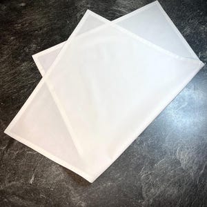Könnte beinhalten: Zwei gefaltete weiße Leinen-Servietten mit dezentem Glanz. Die Servietten sind rechteckig mit einem klaren, minimalistischen Design, perfekt für formelle oder informelle Mahlzeiten. Die Servietten sind auf einer dunklen, strukturierten Oberfläche angeordnet.