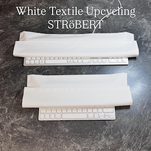 Op de afbeelding: Twee witte textielhoezen geplaatst over twee zilveren computertoetsenborden. De tekst "White Textile Upcycling STRÖBERT" staat bovenaan gedrukt. De hoezen zijn rechthoekig, glad en ondoorzichtig en rusten direct op de toetsen van de apparaten.
