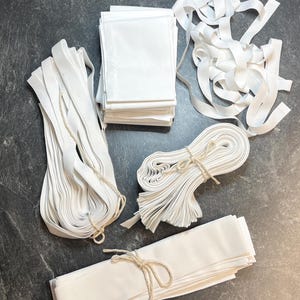 Puede incluir: Una colección de suministros de tela blanca que incluye una pila de piezas rectangulares, un montón de cinta suelta y tres paquetes de tiras de tela asegurados con cordel fino. Los artículos están hechos de material blanco opaco.