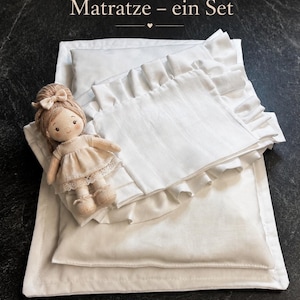 Può includere: Un set di biancheria da letto bianco con una bambola. Il set comprende un cuscino, una coperta e un materasso, tutti bianchi. Il cuscino ha un bordo a balze. Una bambola con un vestito beige e un fiocco è posta sulla biancheria da letto. Il testo nell'immagine recita "Kissen, Decke und Matratze – ein Set" e "für kleine Träume".