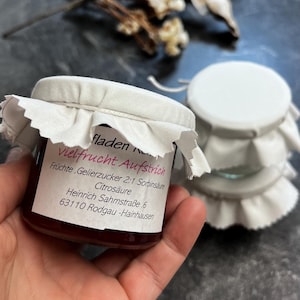 Peut inclure: Un petit pot de confiture de fruits avec un couvercle en tissu blanc et une étiquette avec du texte allemand. Le pot est tenu dans une main, avec deux autres pots empilés en arrière-plan. La confiture est de couleur rouge foncé.