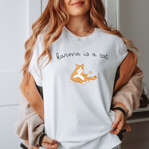 May include: White t-shirt with the text "karma is a cat" and a cartoon orange cat illustration.