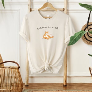 May include: A cream-colored t-shirt with a cartoon orange cat and the text "karma is a cat" printed on the front.