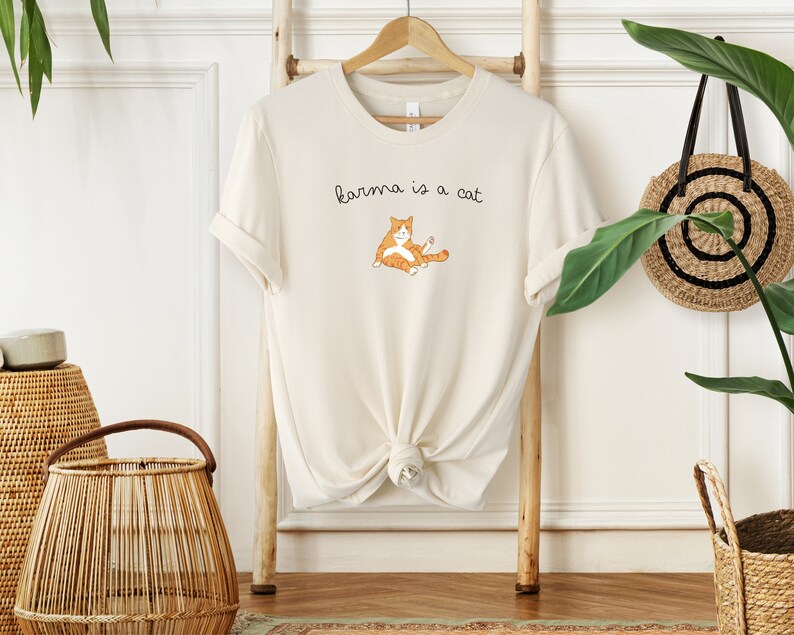 May include: A cream-colored t-shirt with a cartoon orange cat and the text "karma is a cat" printed on the front.