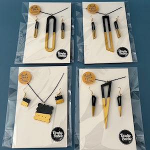 Könnte beinhalten: Vier Sets aus schwarz-goldfarbenem Schmuck, jeweils in einer transparenten Plastiktüte verpackt. Jedes Set enthält eine Halskette und passende Ohrringe. Die Halsketten und Ohrringe haben geometrische Designs. Jedes Paket hat einen kleinen runden Aufkleber mit der Aufschrift "Danke". Der Markenname "Double Bubble" steht auf jeder Verpackung.