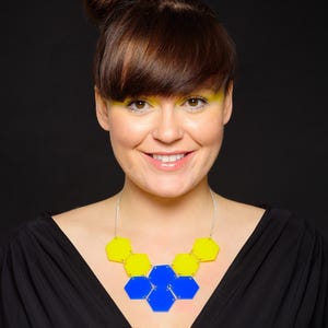 Könnte beinhalten: Eine Frau mit braunen Haaren, die zu einem Knoten gebunden sind, trägt ein schwarzes Oberteil und eine Halskette aus gelben und blauen sechseckigen Formen.
