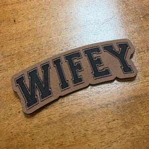 May include: A brown leather patch with the word "WIFEY" in bold, black, block letters. The patch is rectangular with rounded corners and is set against a wooden background.