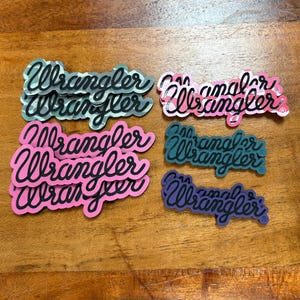 May include: A collection of colorful "Wrangler" logo stickers in various colors, including pink, teal, and purple, with a black outline. The stickers are arranged on a wooden surface.