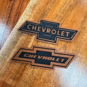 May include: Two Chevrolet logo emblems. The emblems feature a black text and logo design set against a brown background. The word "CHEVROLET" is prominently displayed. The emblems are placed on a wooden surface.