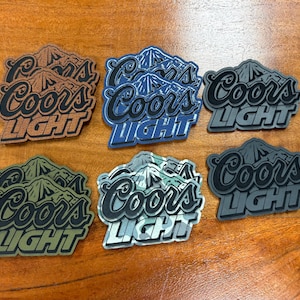 May include: Six Coors Light logo patches in various colors, including brown, blue, gray, green, and camouflage. Each patch features the Coors Light mountain design and text on a wooden surface.