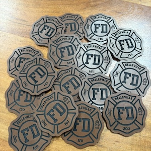 May include: A collection of brown leather patches with a black fire department logo and the text "Belleville FD" and a number. The patches are arranged in a random pattern on a wooden surface.
