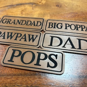 May include: Five rectangular tan leather patches with black borders and black engraved text. The patches feature the words GRANDDAD, BIG POPPA, PAWPAW, DAD, and POPS in a bold serif font. Each patch has a clean, finished edge and a smooth surface.