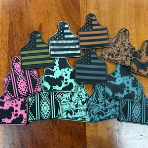 May include: A collection of decorative tags in various designs, including American flag patterns, cowhide prints, and tribal motifs. The tags feature a hole at the top for attachment and are in shades of black, brown, pink, green, and blue.