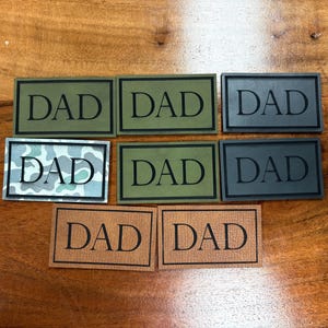 May include: Rectangular patches with the word "DAD" in a bold font. The patches are in various colors, including green, gray, and brown, with black borders. One patch has a camouflage pattern.
