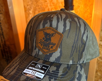 May include: Camouflage trucker hat with a brown and orange patch featuring a deer and the text "Bad Day Turn a Spike." The hat has a mesh back and a curved brim. The tag reads "Richardson Original 112 Trucker."
