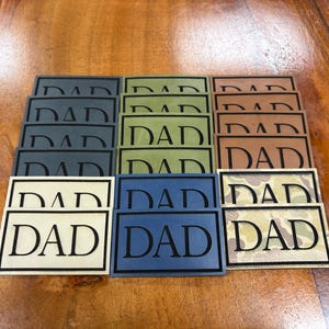 May include: A collection of rectangular patches arranged in rows, each featuring the word DAD in a bold, black serif font. The patches come in various colors including charcoal, olive green, tan, brown, blue, and a camouflage pattern.