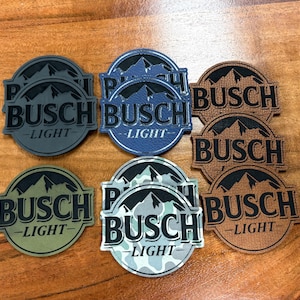 May include: A collection of round patches featuring the "Busch Light" logo in various colors. The patches display a mountain range graphic above the brand name. Colors include gray, blue, brown, green, and camouflage.