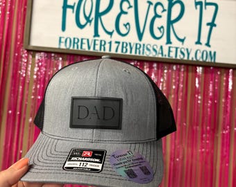 May include: Gray trucker hat with a black mesh back and a black patch that says "DAD". The hat has a Richardson 112 Trucker patch and the text "FOREVER 17" on a sign in the background.