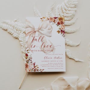 Puede incluir: Una invitación para baby shower con un gran lazo y detalles florales. La invitación dice "Fall in love with Baby Hudson" e incluye detalles del evento. Colores suaves y neutros, diseño elegante.