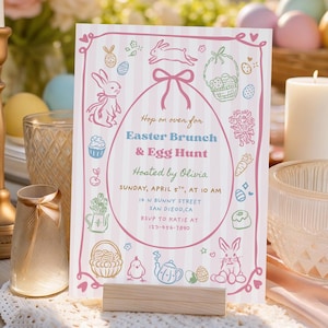 Puede incluir: Una tarjeta de invitación a un brunch de Pascua y una búsqueda de huevos sobre un soporte de madera. La tarjeta tiene un fondo a rayas rosas y blancas con ilustraciones y texto con temática de Pascua. La invitación está colocada sobre una mesa con velas, flores y huevos de Pascua.