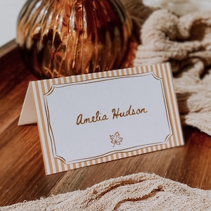 May include: A place card with the name "Amelia Hudson" in gold script, featuring a leaf emblem. The card has a white background with gold stripes and a decorative gold border. A brown glass vase and dried plants are in the background.