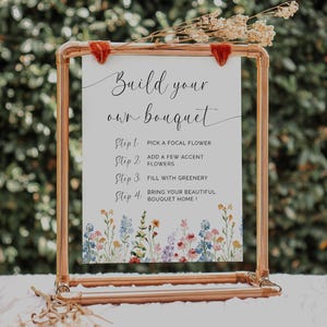 Puede incluir: Un marco de metal color cobre sostiene un letrero blanco con el texto "Build your own bouquet". El letrero enumera los pasos para crear un ramo, con un borde floral. Flores secas se colocan en la parte superior.