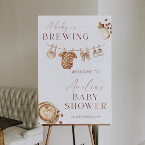 May include: A baby shower sign on a wooden easel. The sign displays the text "A baby is brewing" and "Welcome to Amelia's Baby Shower" in a stylish font. The sign also features illustrations of baby clothes and a coffee cup.