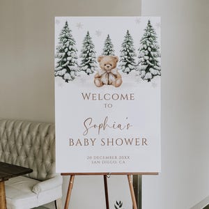 May include: A white baby shower welcome sign on a wooden easel. The sign features a teddy bear, snow-covered pine trees, and the text "Welcome to Sophia's Baby Shower" with the date and location.