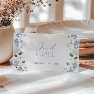 Könnte beinhalten: Ein weißes Lebensmitteletikett mit blauen floralen Akzenten und den Worten "Food Label" in eleganter Schrift. Das Etikett befindet sich auf einem Tisch mit Tellern und einer Vase mit Trockenblumen, was eine rustikale, elegante Ästhetik schafft.