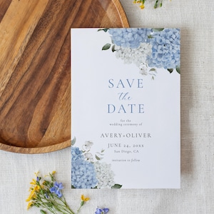 May include: A white wedding invitation with blue floral accents and the text "SAVE THE DATE". The invitation includes the names AVERY + OLIVER, the date June 24, 20XX, and the location San Diego, CA. A wooden tray and flowers are also in the image.