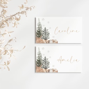 Puede incluir: Dos tarjetas rectangulares blancas con texto dorado e ilustraciones de acuarela. Cada tarjeta presenta un &aacute;rbol de Navidad en una cesta tejida, regalos y copos de nieve. Una tarjeta dice "Caroline" y la otra "Amelia".