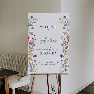 May include: A white welcome sign with a floral border and the text "Welcome to Amelia's bridal shower 21.08.2027 San Diego, California".