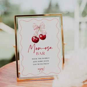May include: A printable sign for a mimosa bar featuring a pink bow and two red cherries. The sign reads "Mom-osa Bar" and includes instructions for making mimosas: "Pour the bubbly, add some juice, top with fruit, enjoy!"