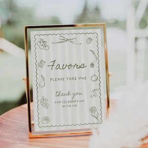 K&ouml;nnte beinhalten: Ein goldgerahmtes Schild mit dem Wort "Favors" in Schrift, umgeben von einem gewellten Rand mit Fruchtdarstellungen. Das Schild lautet "Please take one" und "thank you for celebrating with us!" auf gestreiftem Hintergrund.