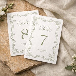 May include: Two white rectangular table number cards featuring a sage green floral vine border, the word Table in script, the numbers 7 and 8, and the text Grace & Nathan 10.23.20XX. The cards have a textured paper finish and elegant botanical illustrations.
