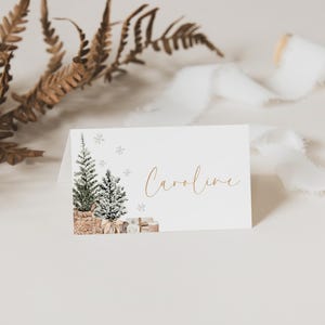 Puede incluir: Tarjeta blanca plegada con una ilustraci&oacute;n de acuarela de dos &aacute;rboles de Navidad, copos de nieve y regalos. El nombre "Caroline" est&aacute; escrito en letras doradas. Hojas secas y cinta blanca en el fondo.