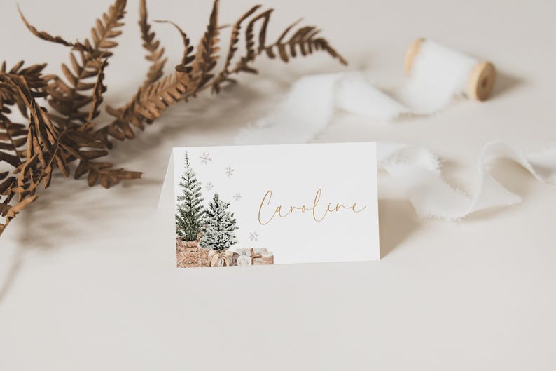 Puede incluir: Tarjeta blanca plegada con una ilustraci&oacute;n de acuarela de dos &aacute;rboles de Navidad, copos de nieve y regalos. El nombre "Caroline" est&aacute; escrito en letras doradas. Hojas secas y cinta blanca en el fondo.