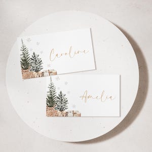 Puede incluir: Dos tarjetas blancas con los nombres "Caroline" y "Amelia" en escritura dorada. Cada tarjeta presenta una ilustraci&oacute;n en acuarela de un &aacute;rbol de Navidad, regalos y copos de nieve. Las tarjetas est&aacute;n sobre una superficie circular blanca.