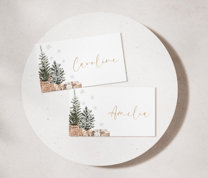 Puede incluir: Dos tarjetas blancas con los nombres "Caroline" y "Amelia" en escritura dorada. Cada tarjeta presenta una ilustraci&oacute;n en acuarela de un &aacute;rbol de Navidad, regalos y copos de nieve. Las tarjetas est&aacute;n sobre una superficie circular blanca.