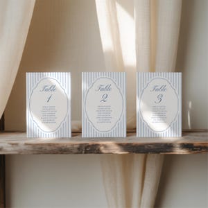 May include: Three rectangular table cards with a blue and white striped background. Each card has a decorative oval frame with the word "Table" and a number. Below the frame are names. The cards are on a wooden surface.