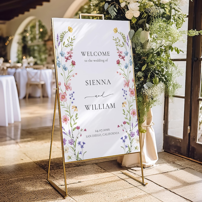 Puede incluir: Un letrero de bienvenida de boda con marco dorado y detalles florales. El letrero dice "Bienvenida a la boda de Sienna y William" con la fecha y el lugar. El letrero est&aacute; decorado con flores de acuarela en rosa, azul, amarillo y morado.
