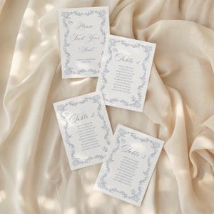 May include: Four rectangular white wedding seating cards with blue floral borders. The top card reads Please Find Your Seat, Grace & Nathan, 10.25.20XX. The other three cards are labeled Table 1, Table 2, and Table 3, each listing guest names.