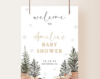 May include: Welcome to Amelia's Baby Shower sign with a winter theme. The sign features a white background with gold lettering and illustrations of snow-covered pine trees and a small wooden house. The text on the sign reads "Welcome to Amelia's Baby Shower 12.14.25 San Diego, CA."