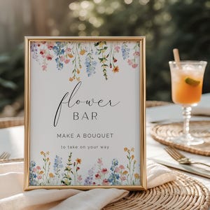 May include: A framed sign with a gold border, featuring the words "Flower Bar" and "Make a Bouquet to take on your way." The sign is decorated with watercolor flowers in shades of pink, blue, yellow, and purple. A cocktail sits in the background.