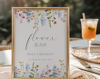 May include: A framed sign with a gold border, featuring the words "Flower Bar" and "Make a Bouquet to take on your way." The sign is decorated with watercolor flowers in shades of pink, blue, yellow, and purple. A cocktail sits in the background.